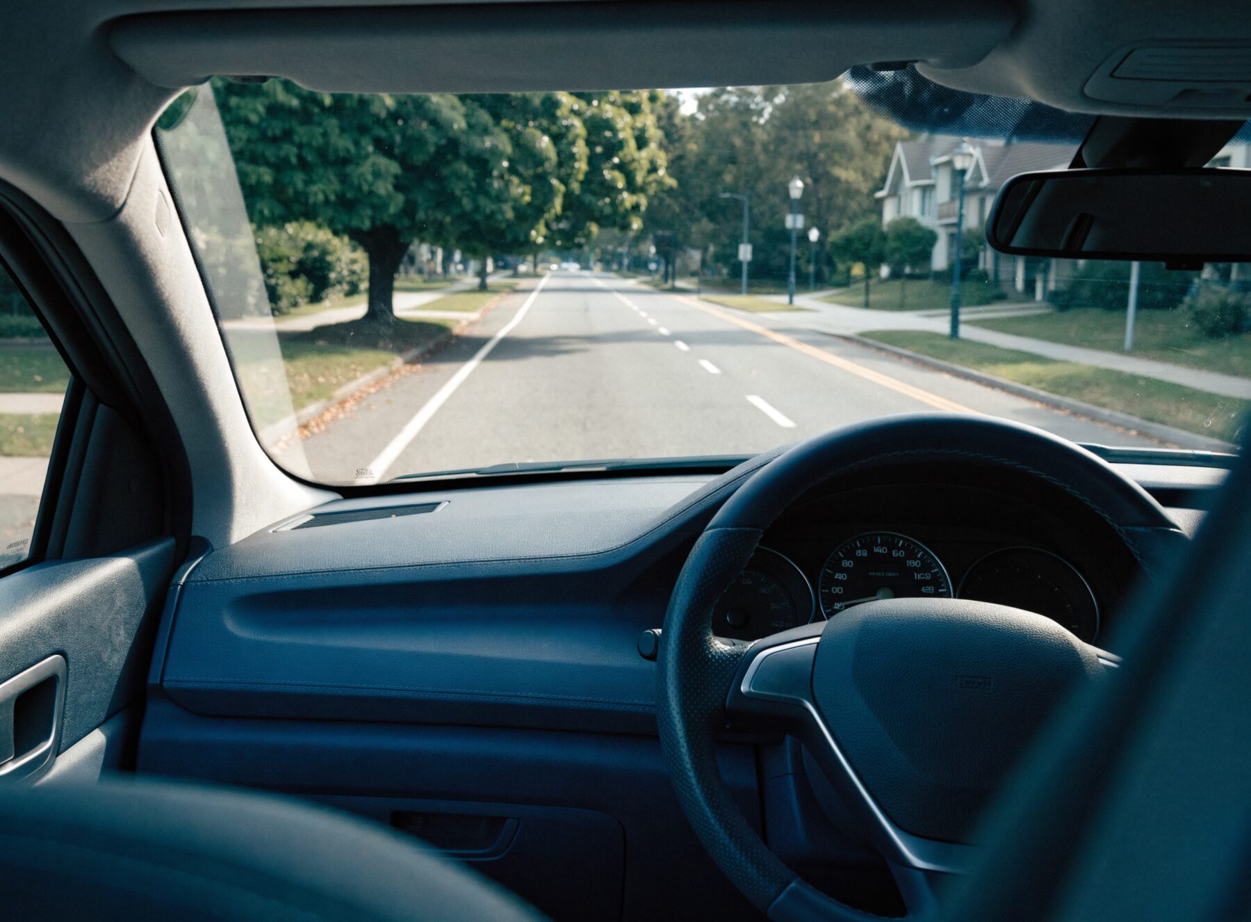 View across the dashboard and steering wheel of a manual driving school car on a quiet tree-lined street